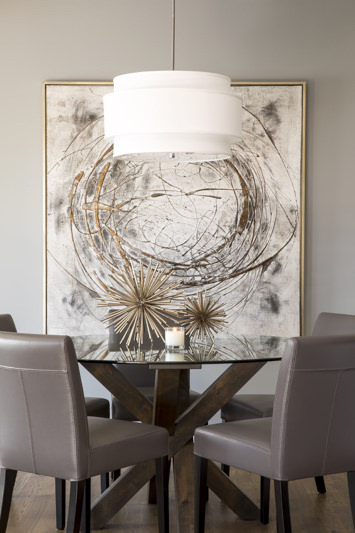 Modern dining area with a glass table, gray chairs, a white pendant light, abstract art, and a lit candle. Neutral tones create a calm mood.