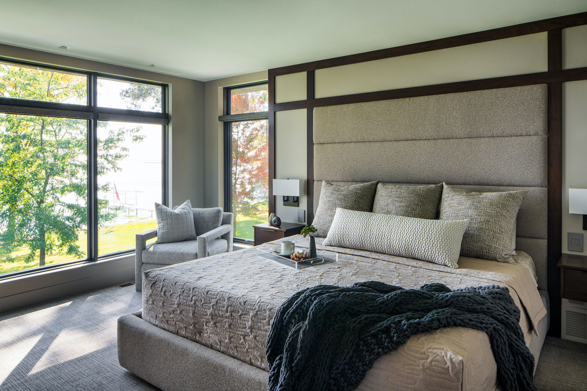 Modern bedroom with large windows showing a scenic view. Gray bed with textured bedding, cozy pillows, and a plush chair create a serene vibe.