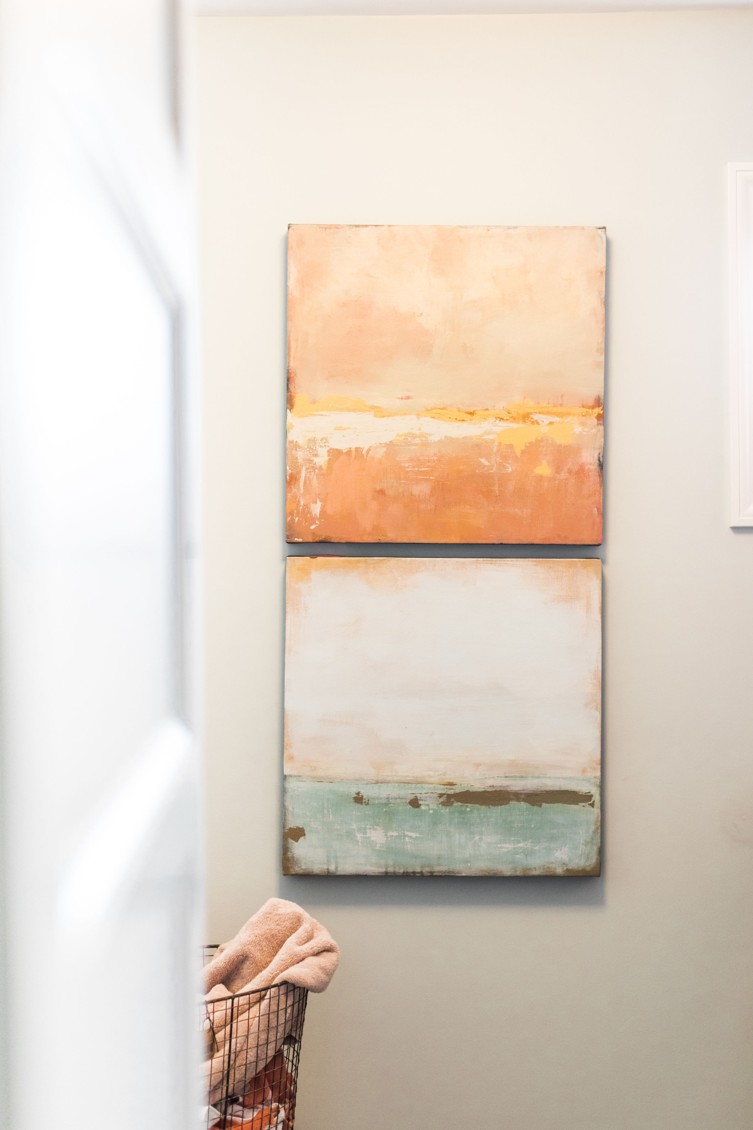 Two abstract paintings in peach and teal tones hang on a light wall. A wire basket with a pink towel is partially visible below. Calm setting.
