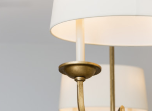 Close-up of a brass chandelier, featuring white lampshades and a candle-shaped bulb, set against a soft, neutral background.