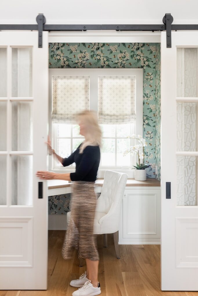 Woman slides white barn door in floral office with sunlight through window, white chair and green wallpaper. Bright, airy atmosphere.