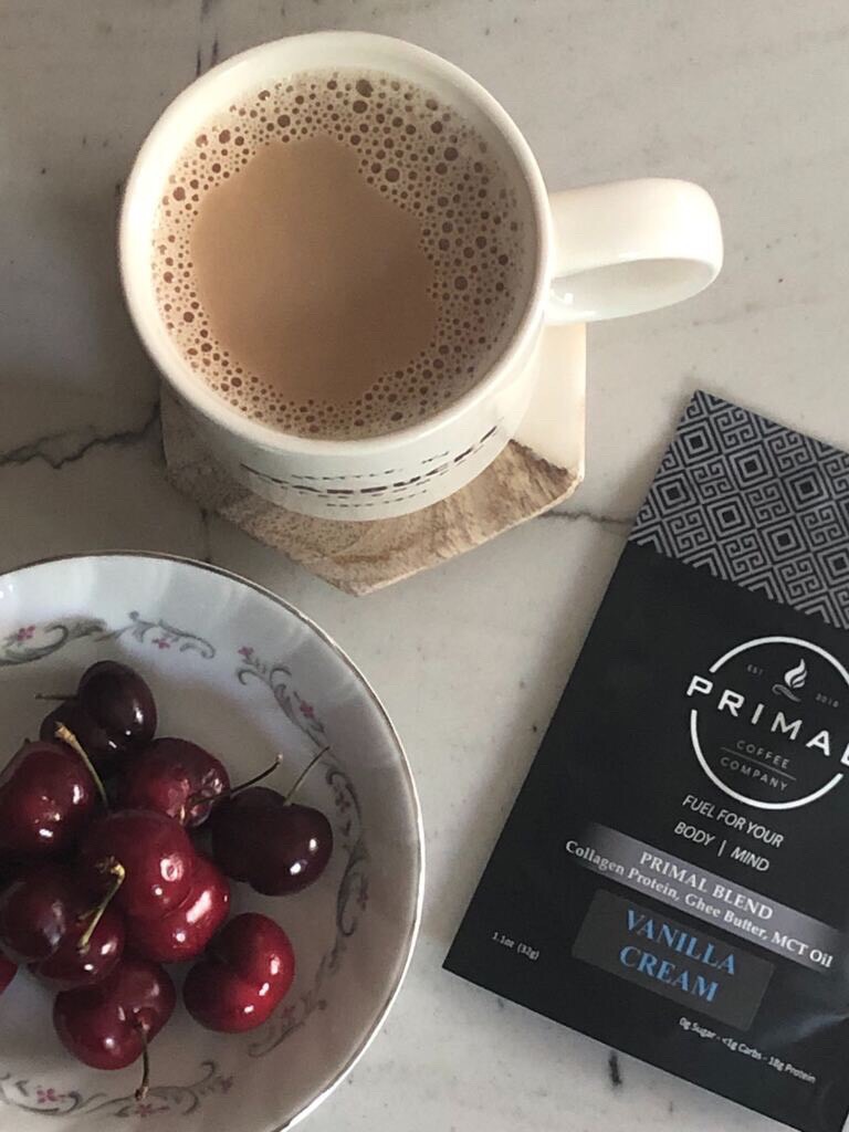 A white mug of coffee on a coaster, next to a bowl of cherries and a black Primal Coffee packet labeled "Vanilla Cream" on a marble surface.