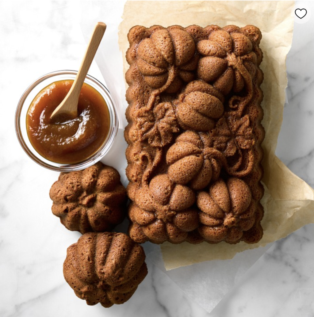 Brown pumpkin-shaped cake on parchment paper, with two small bundt cakes and a jar of caramel sauce with a wooden spoon on a marble surface.