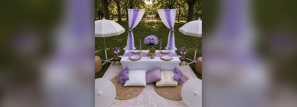 Luxe Birthday Picnic by The Picnic Loft in Charlotte, NC, with lavender and white draped arch, Happy Birthday neon sign, white fringe umbrellas, purple floral centerpiece, and lavender floor pillows on a park lawn.
