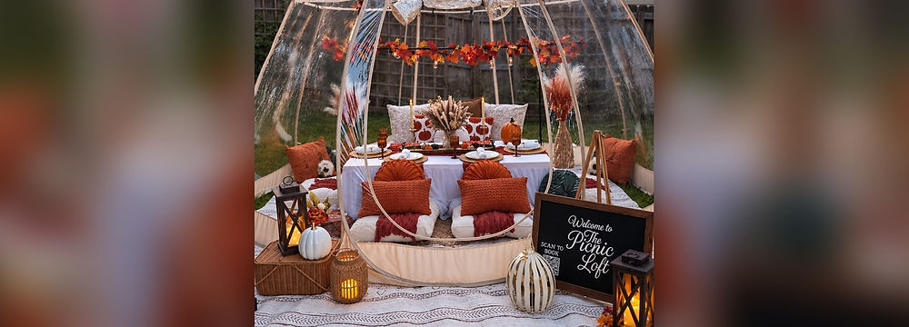 The Harvest Glow fall globe tent picnic by The Picnic Loft in Charlotte, NC, with an autumn leaf garland and string lights, pampas grass, pumpkins, amber glassware, and rust orange floor pillows around a low table.