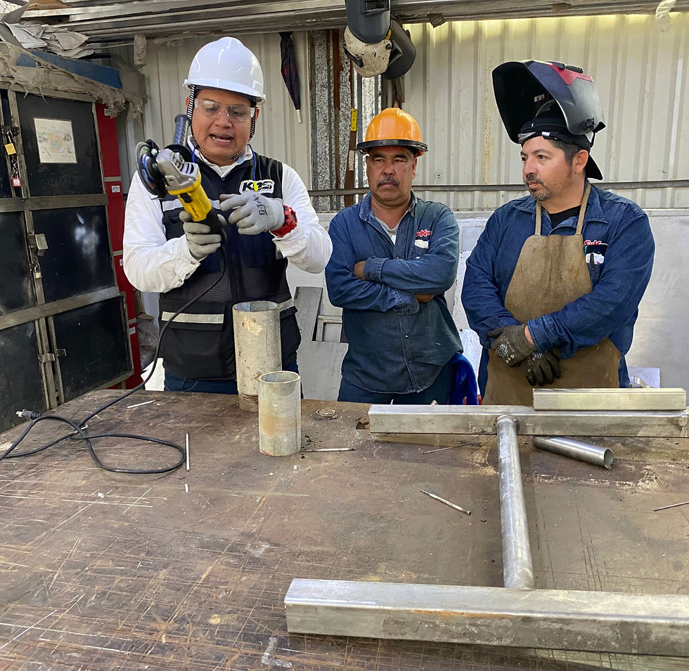 Tres hombres en un taller, uno con casco blanco explica el uso de una herramienta. Los demás observan, uno con casco naranja y otro con soldadura.