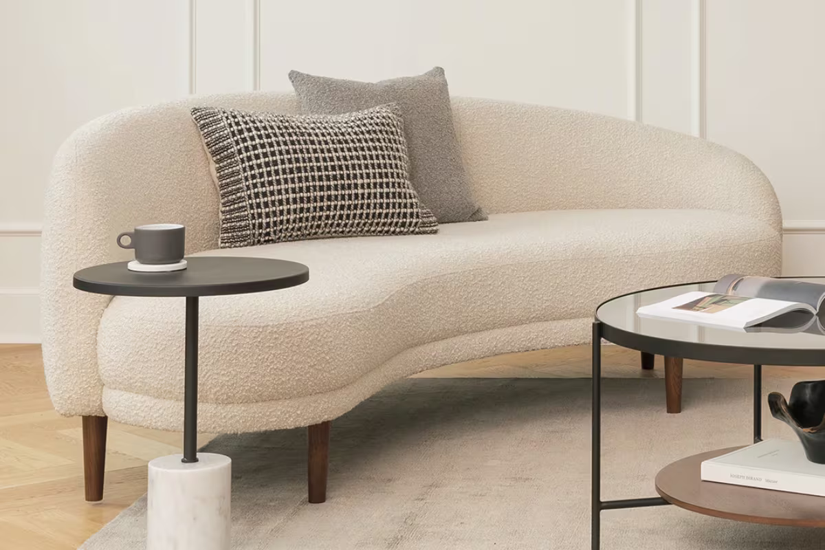 Beige curved sofa with black printed and textured cushions and a medium gray square cushion with a black top side table with white marble base