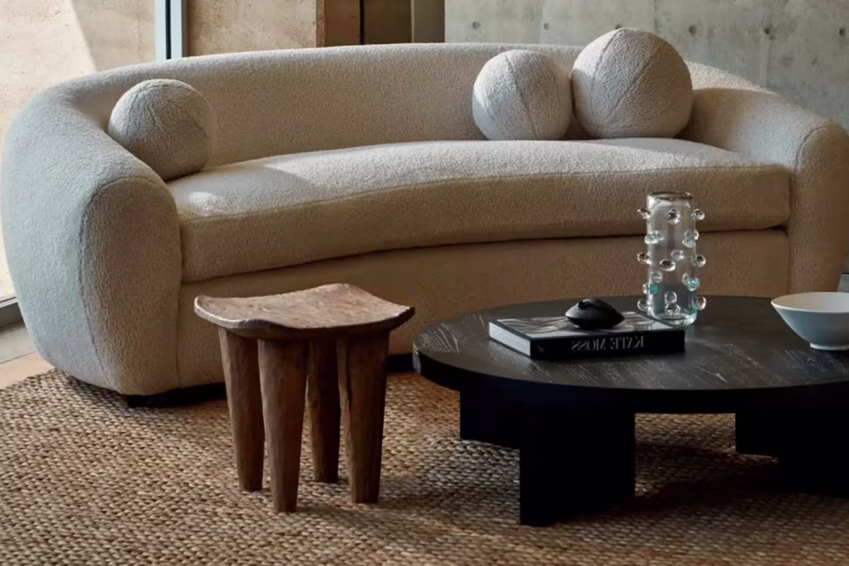 Beige textured fabric sofa with round beige cushions and a black coffee table made of wood in black lacquer and a wooden stool all placed on a textured brown carpet