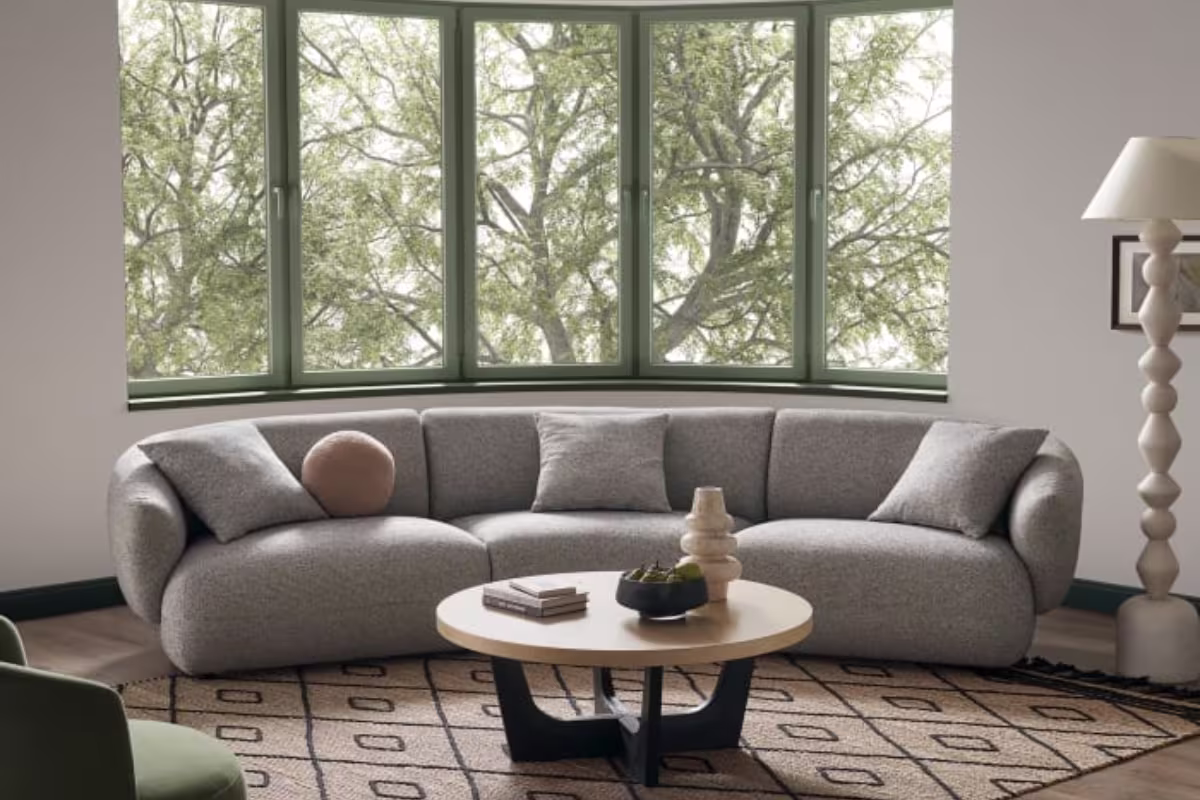 Gray curved sofa with gray cushions placed in a corner with a curved window with a wooden top coffee table with black base and a printed textured carpet