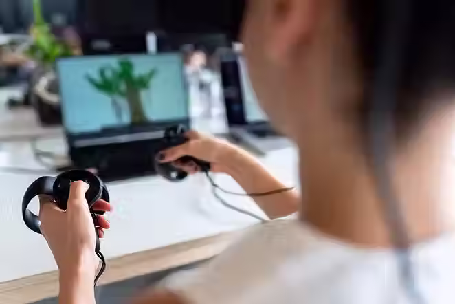 Person wearing a headset uses VR controllers at a desk, with a laptop showing a plant scene in a bright home office.