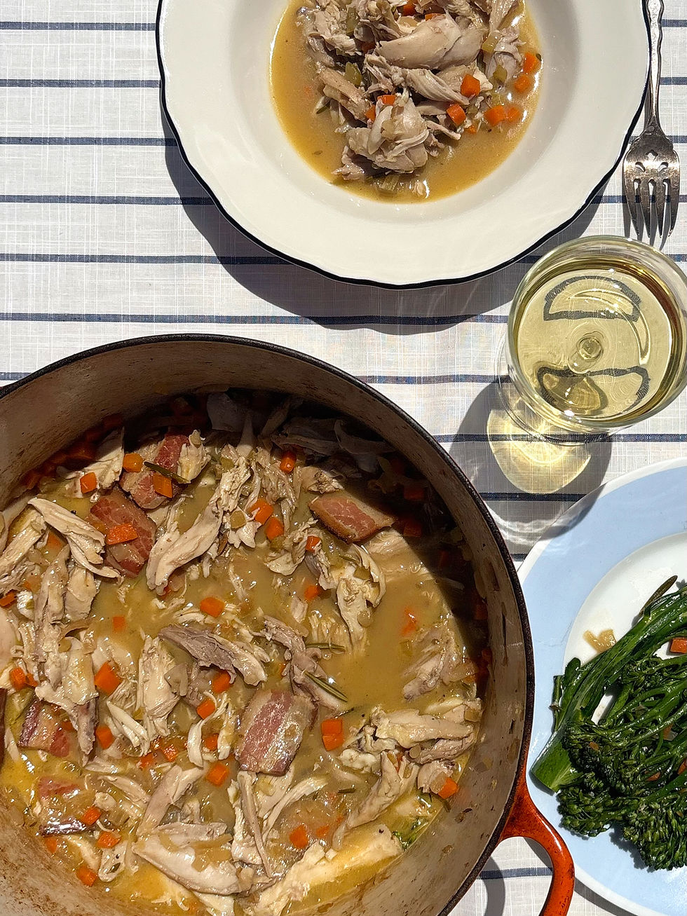 Braised chicken and vegetables in a pot next to a served plate on a blue-striped tablecloth. Broccolini and a glass of white wine nearby. French stewed chicken. Countryside cottage core meal. Wholesome food that is nourishing and hearty. Summer dinner. Winter dinner. Everyday meal prep. Chicken stew. white wine chicken. Dutch oven braised chicken.