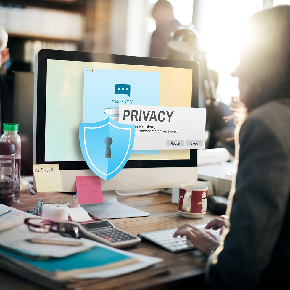 Person typing on a keyboard in an office. A computer screen shows a privacy warning and a blue shield icon. Sunlight filters through a window.