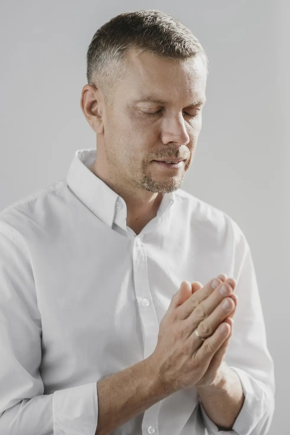 Middle-aged man in contemplative prayer, eyes closed and hands joined in a quiet indoor setting.