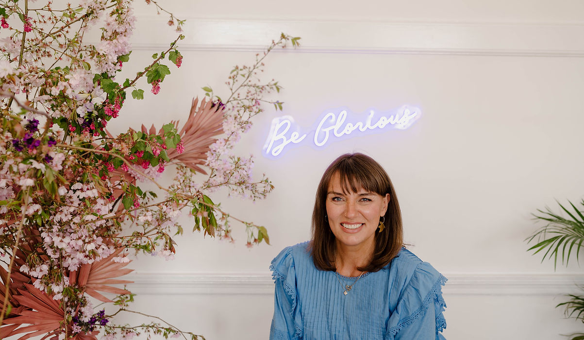A woman with mid length dark blonde hair wearing a denim blue top with frilled broderie anglaise shoulders and a neon sign on the wall behind her which reads 'Be Glorious.' The woman is UK based wedding accessory designer Heidi Reid of Glorious by Heidi.