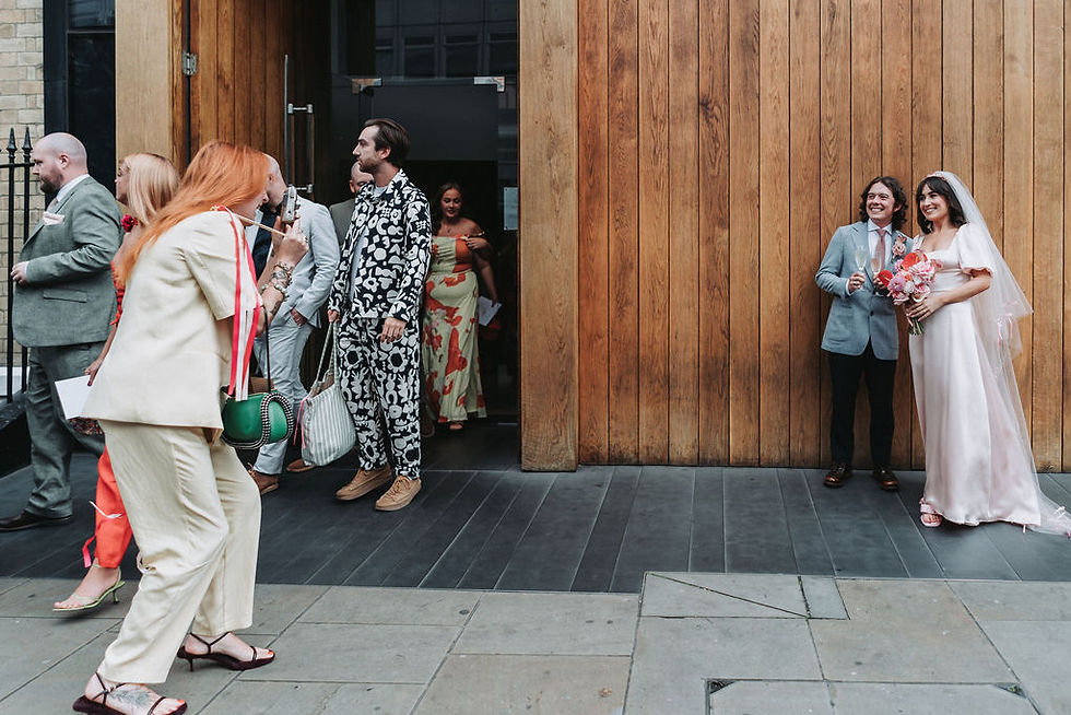 Bride in pink dress and pink crystal crown and groom with champagne smile by wooden wall, guests exit building on left.