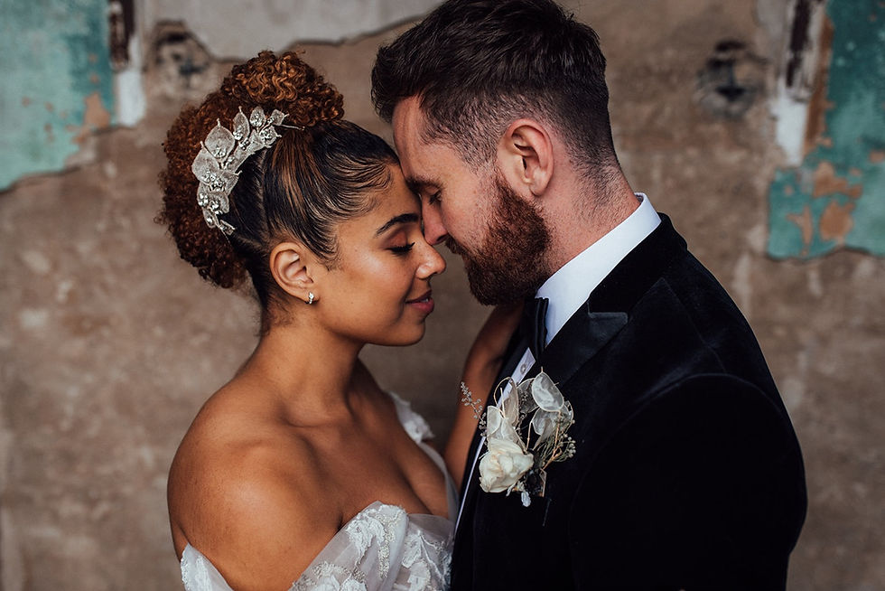 Thumbnail: Image shows the head and shoulders of a male and female model posing as a newly wed couple with their foreheads touching. The bride is black and has afro hair styled in a mohawk hairstyle. Her hairstyle is accessorised with a duo of silver leaves and crystal buds wedding accessories pinned either side of her head. The groom is white and has a full beard. He wears a velvet jacket and copper coloured tie.