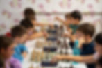 Children play chess at long tables in a bright room with floral wallpaper, focused hands moving pieces over digital chess clocks.