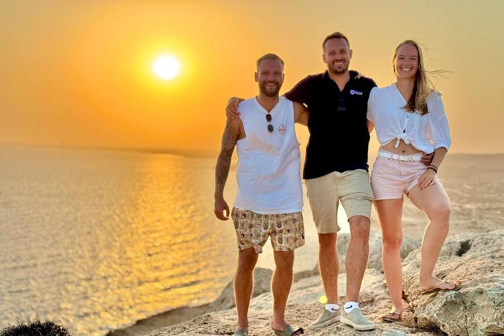 Drei Personen (Joel, Daniel und Tamara) stehen lächelnd bei Sonnenuntergang an einer felsigen Küste. Im Hintergrund glitzert das Meer im goldenen Licht der untergehenden Sonne, der Himmel ist in warmen Gelb- und Orangetönen gehalten. Sie tragen leichte Sommerkleidung: Joel ein weisses Tanktop und Shorts, Daniel ein dunkles Poloshirt und Shorts, Tamara ein weisses Oberteil und kurze helle Shorts.