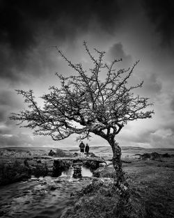 A black and white photo of a couple on a stone bridge across a stream, framed by a tree and surrounded by a moody Dartmoor landscape