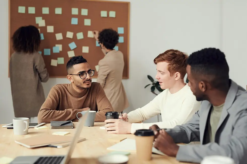 A team in the office. 2 women are putting sticky notes on the wall board and 3 men are sitting at the table, having a chat over coffee.