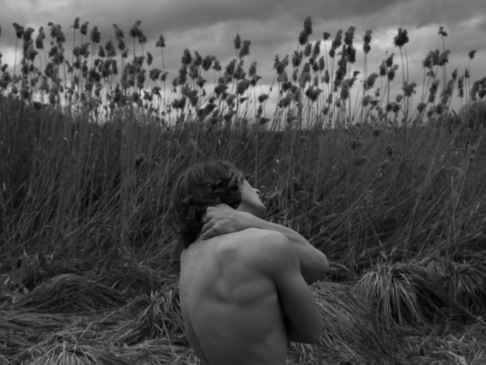 Shirtless person with long hair holds neck in a grassy field under moody, cloudy sky. Tall plants sway in the background. Black and white image.