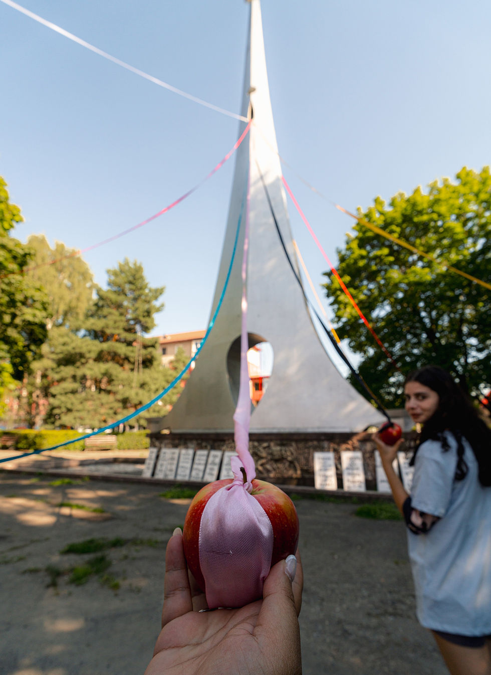 Modern sculpture wrapped in colorful ribbons: red, yellow, blue; set against a sunny blue sky and green trees, creating a vibrant scene.