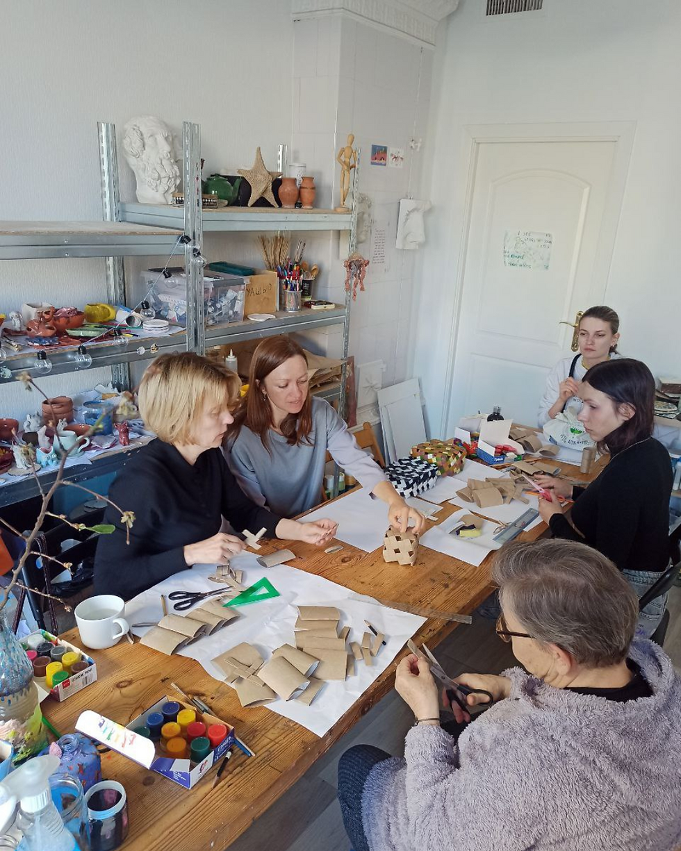 Five people craft with paper at a wooden table in a cozy room. Shelves hold art supplies. The mood is creative and focused.