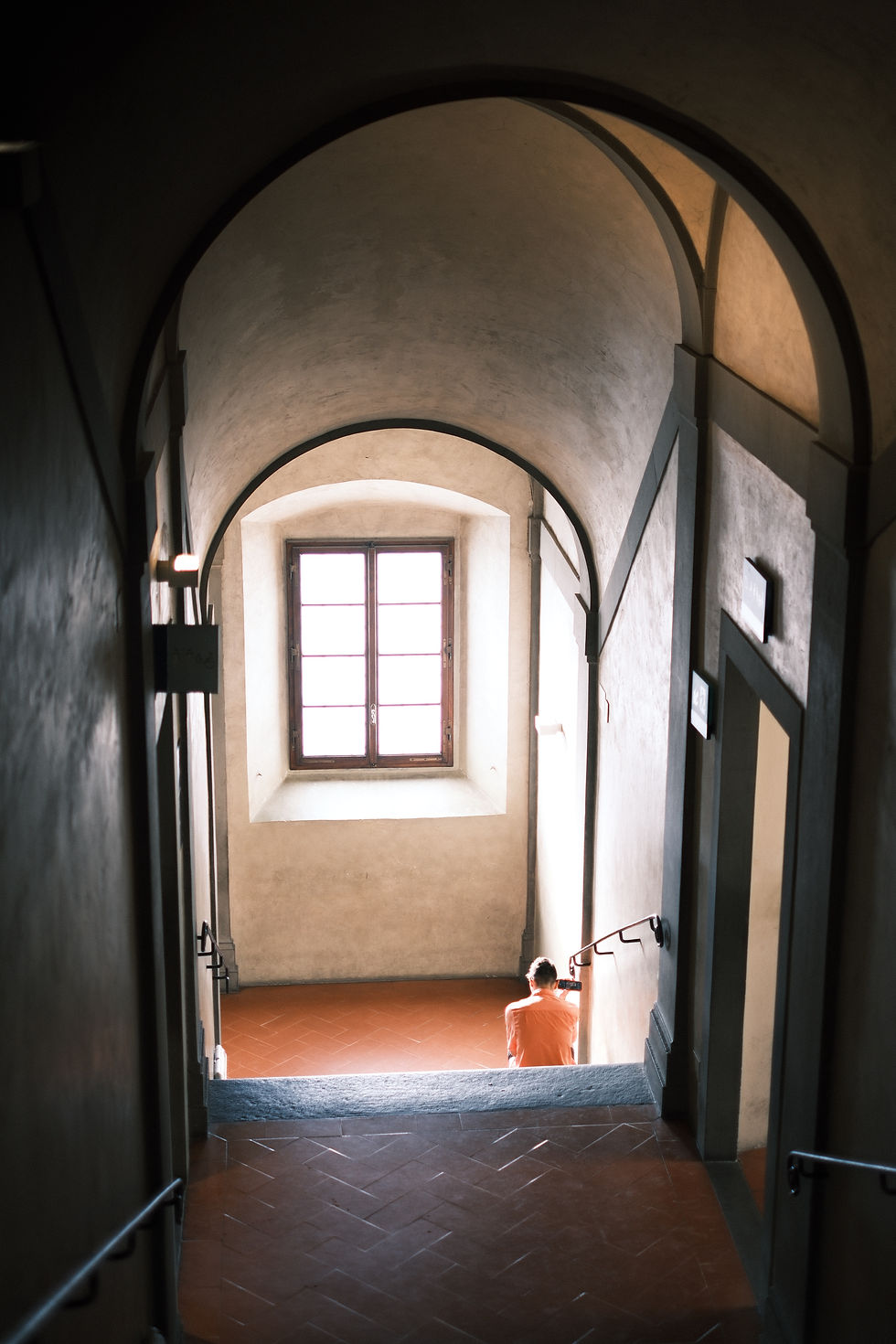 Person in an orange shirt sits on a stair landing in a dim arched hallway, lit by a bright window.
