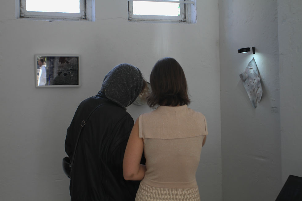 Two people stand in an art gallery, observing a piece on a white wall. One wears a hood, the other a beige top. Natural light shines in.