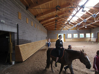 Lana during a hippotherapy session while riding a horse in an indoor riding arena with a physiotherapist leading the horse.