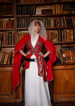 Lana Salome Kurasidze stands confidently in a traditional Georgian costume with a long white dress, red cape and matching accessories, posing in front of bookshelves in a historic library while using a walker for support.