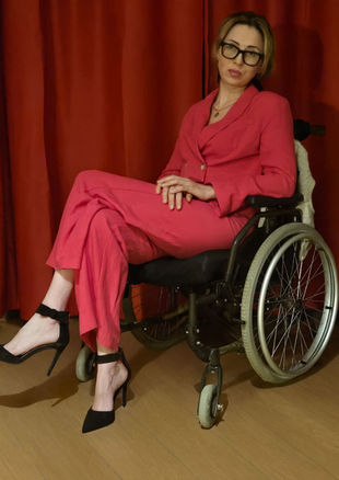 Lana Salome Kurasidze sits confidently in a wheelchair wearing a fuchsia trouser suit, black high heels and glasses, with her hair tied back, posing with a strong and confident expression.