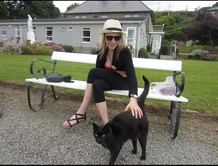 Lana sitting on a white bench, smiling while petting a black cat, wearing sunglasses.