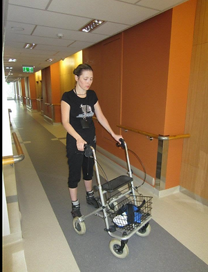 Lana walking with a rollator through a rehabilitation centre corridor while wearing sports clothing.