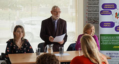 Lana and another BADIG committee member seated at a conference table while the former Chairperson delivers a speech to the audience.