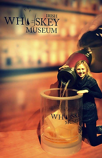 Lana kneeling beside a giant whisky bottle and a large whisky glass display at the Irish Whiskey Museum, smiling at the camera.