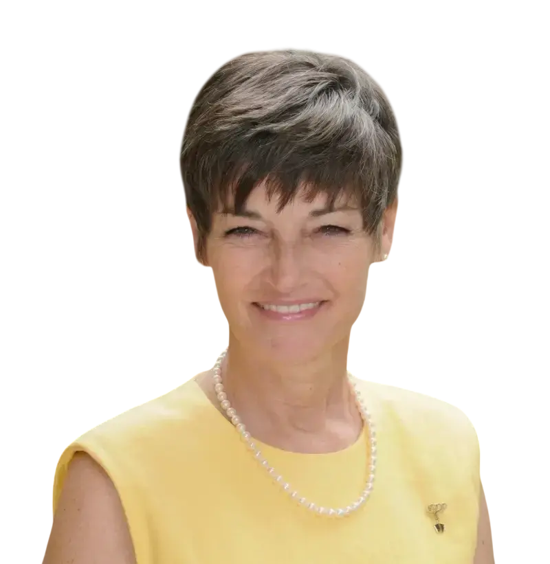 Studio portrait of a middle-aged woman with short hair wearing a yellow top and a pearl necklace. She is smiling warmly against a solid black background, conveying a professional and friendly demeanor.