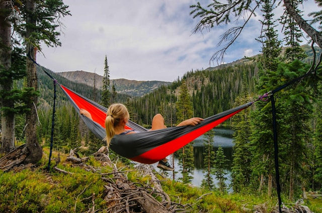 Person relaxing in a red hammock, surrounded by pine trees with a lake and mountains in the background under a partly cloudy sky.