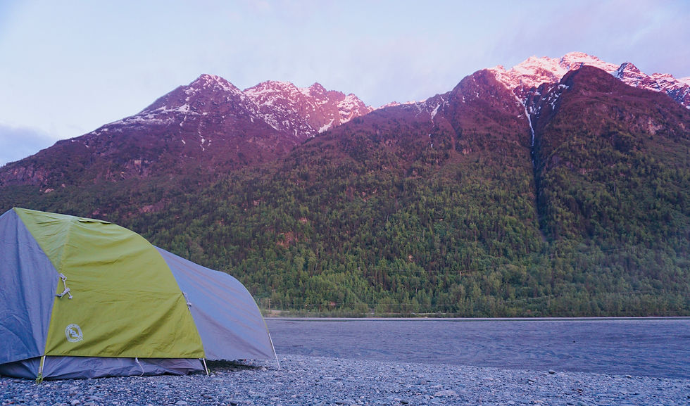 Green and gray tent on rocky shore in front of lush, tree-covered mountains with snowy peaks at sunrise. Peaceful and scenic setting.