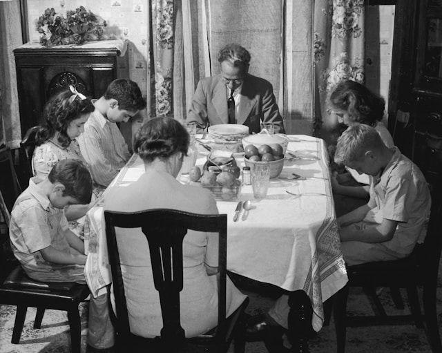 Family of seven bows heads in prayer around dining table. Black and white image, vintage decor, floral curtains, and fruit bowl visible.