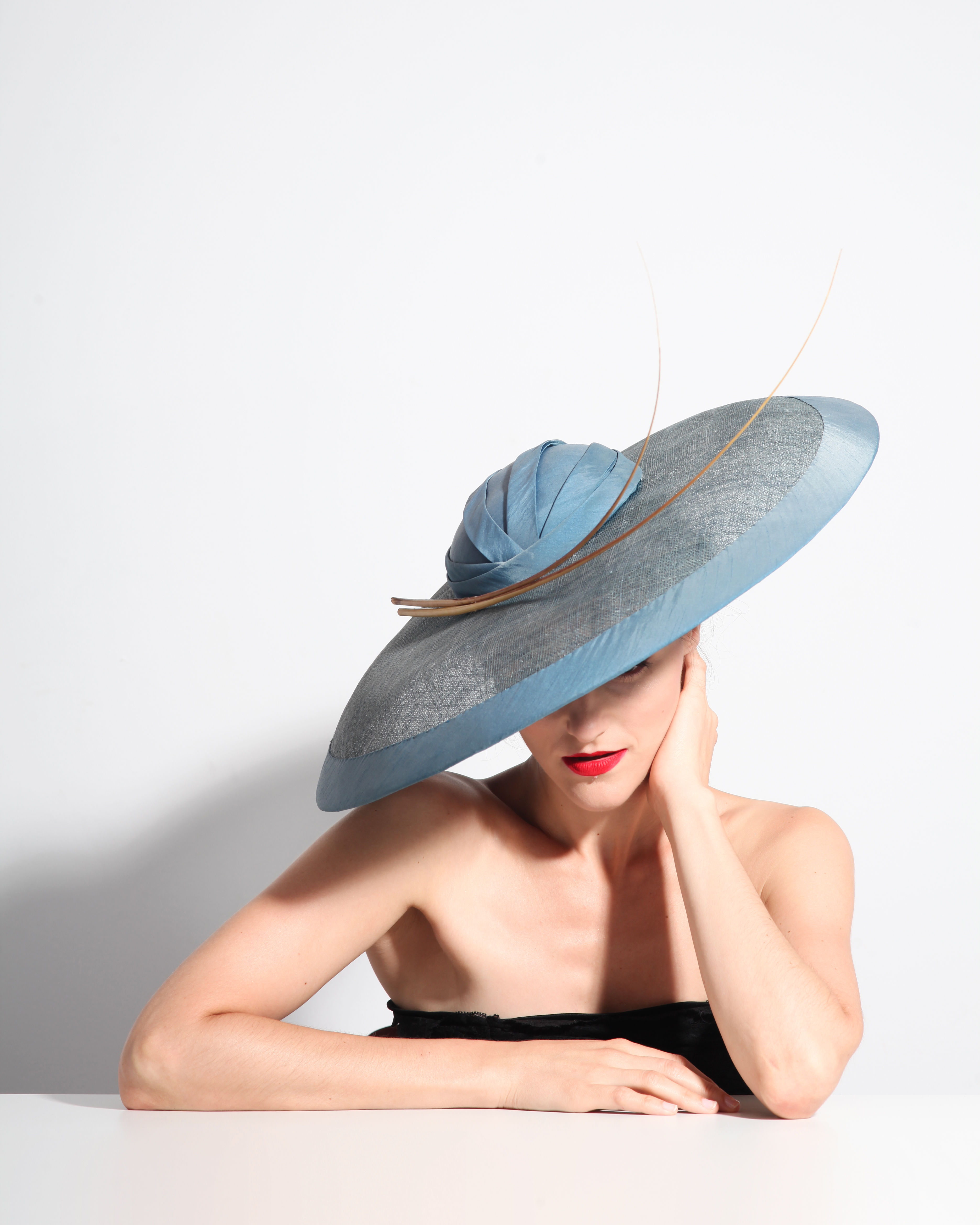 Grand chapeau bleu en sinamay fait main dans l’atelier Maison Eremina à Paris, à bords plats, orné de longues plumes fines.