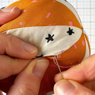 Hands use a needle and orange thread to make a blind stitch to close the opening of a fabric orange and white basketball