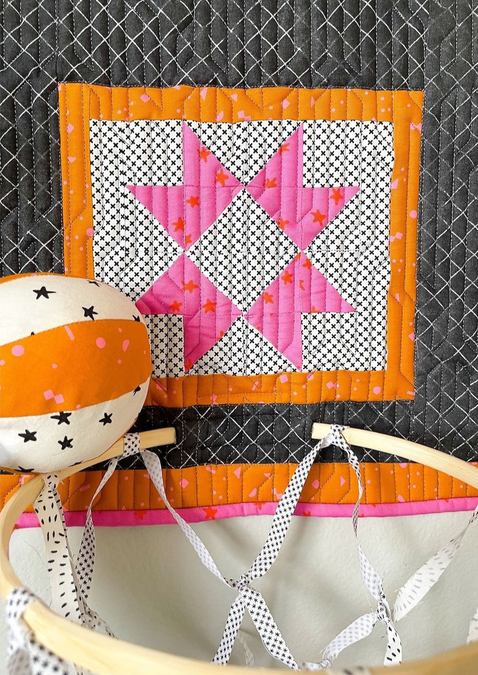 A white and orange fabric basketball sits on the wooden rim of a black, orange, white and pink fabric basketball hoop