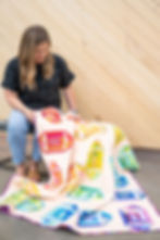 A woman in a black shirt and jeans looks at a rainbow colored scrappy football quilt laying on her lap draping to the concrete floor.