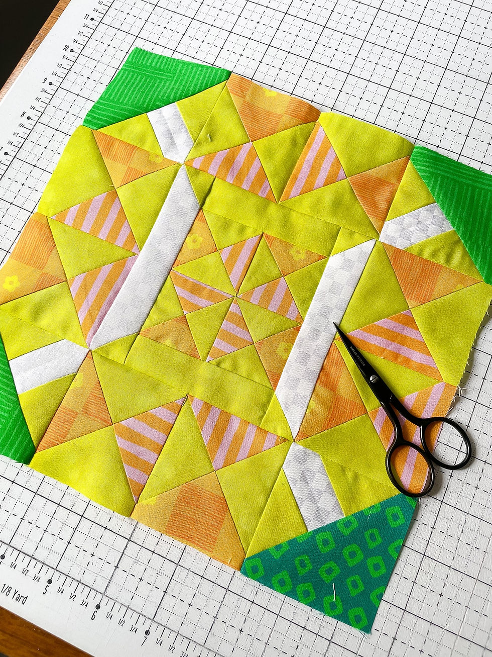 A pieced tennis ball quilt block made of yellow, orange and pink fabrics sits on a white cutting mat with a small pair of black scissors sitting on top.