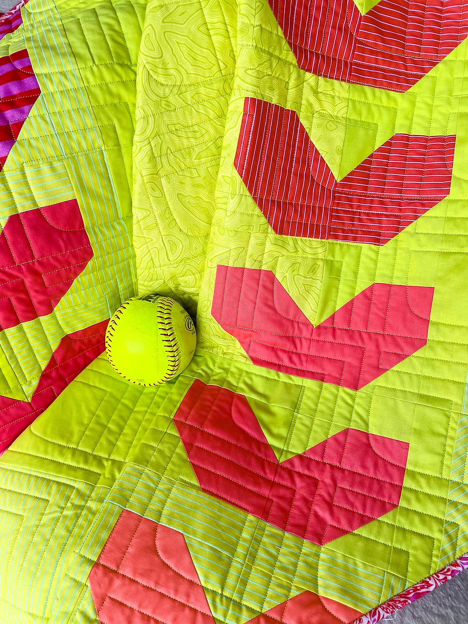 A softball laying on top of a quilt that looks like a softball in neon yellow and red colors
