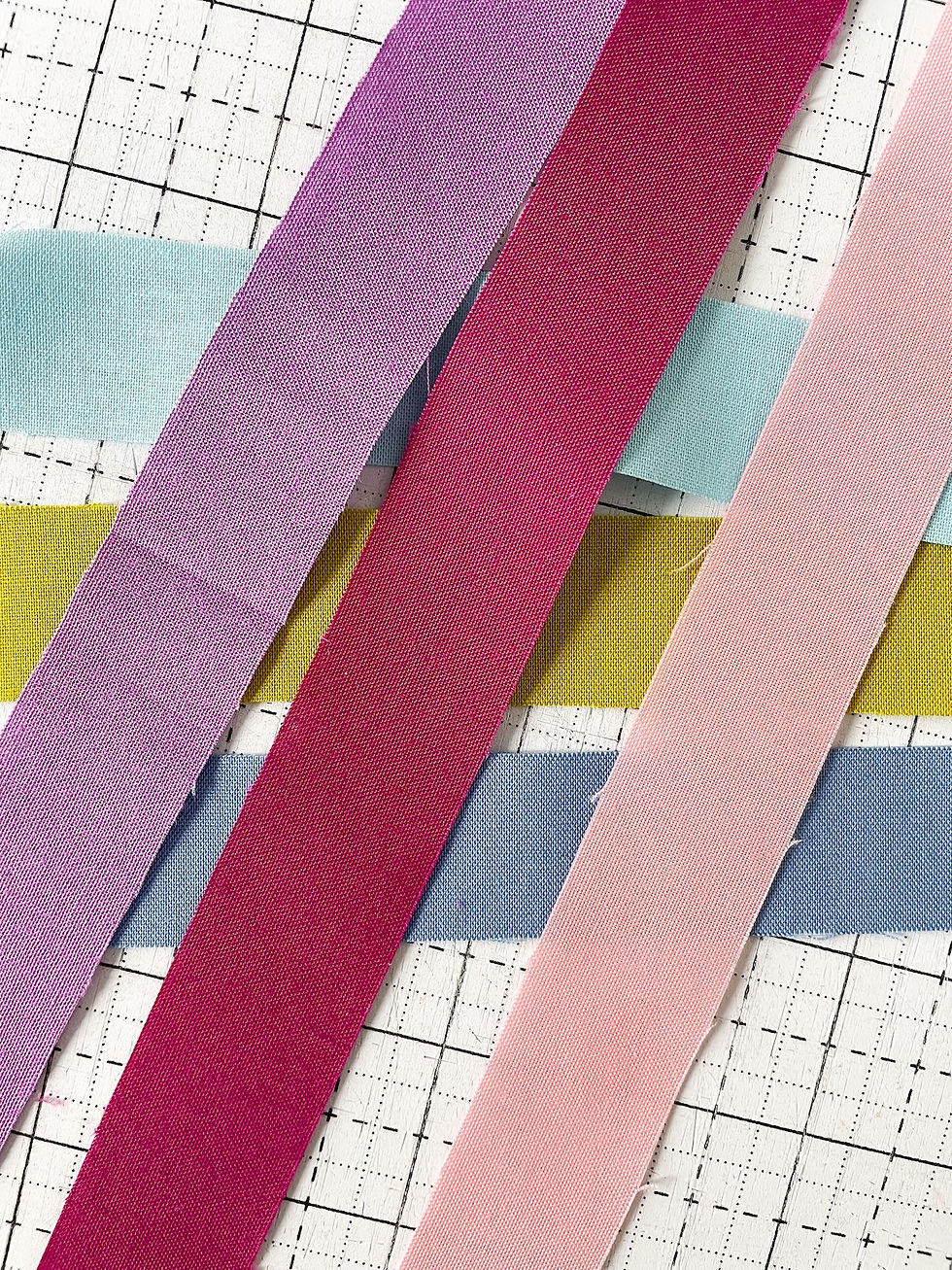 Strips of purple, blue and green fabric lay crossed on a white sewing cutting mat.