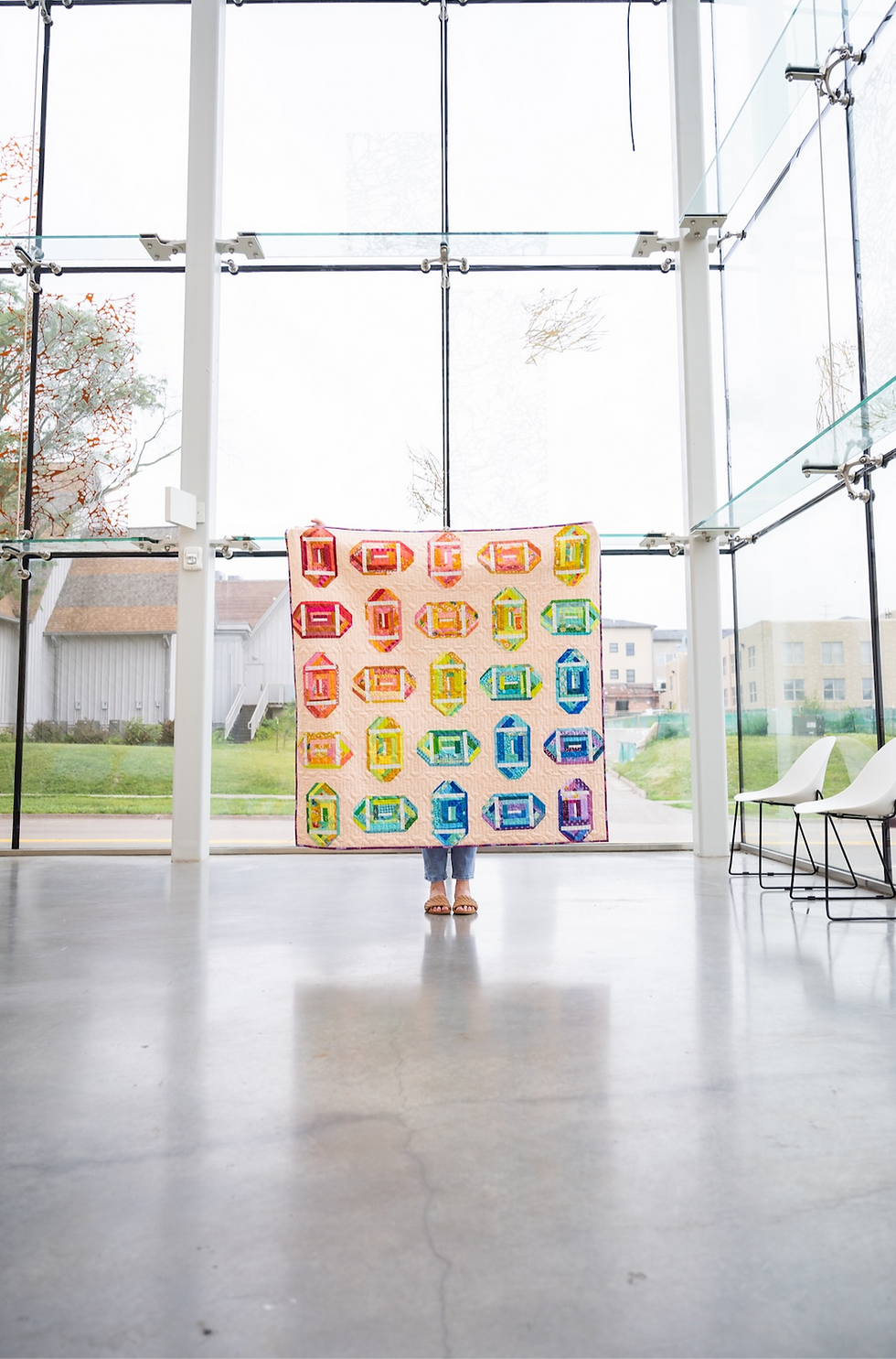 A person holds a rainbow colored football quilt made from scraps of fabric with a neutral background standing on a concrete floor.