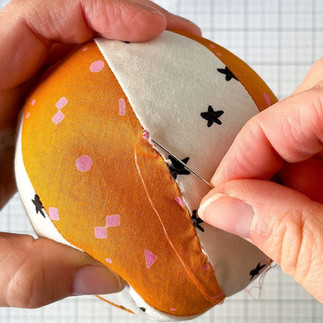 Hands use a needle and orange thread to make a secure knot to close the opening of a fabric orange and white basketball.