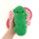 A hand holds a smiling green plush pickle toy with yellow cheeks. A red checkered pattern fry basket in the background. Bright and cheerful.